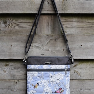 Pu&ograve; includere: Una borsa a tracolla grigia e blu con stampa floreale e farfalle. La borsa ha due tasche con cerniera e una lunga tracolla regolabile.