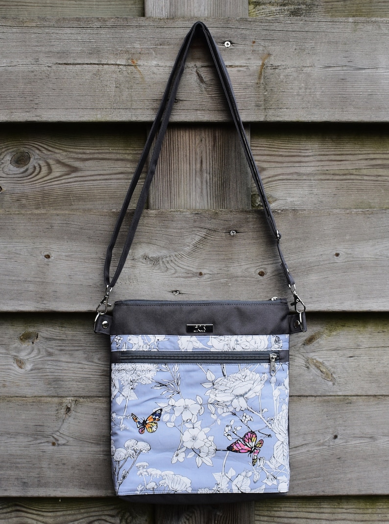 Pu&ograve; includere: Una borsa a tracolla grigia e blu con stampa floreale e farfalle. La borsa ha due tasche con cerniera e una lunga tracolla regolabile.