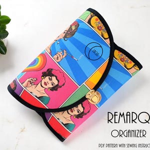 May include: A colorful organizer with a pop art comic book design. The cover features panels with illustrations of women, a rainbow, and the text "LACUS". The organizer has a black trim and a gold clasp. The text "REMARQ ORGANIZER" is also visible.