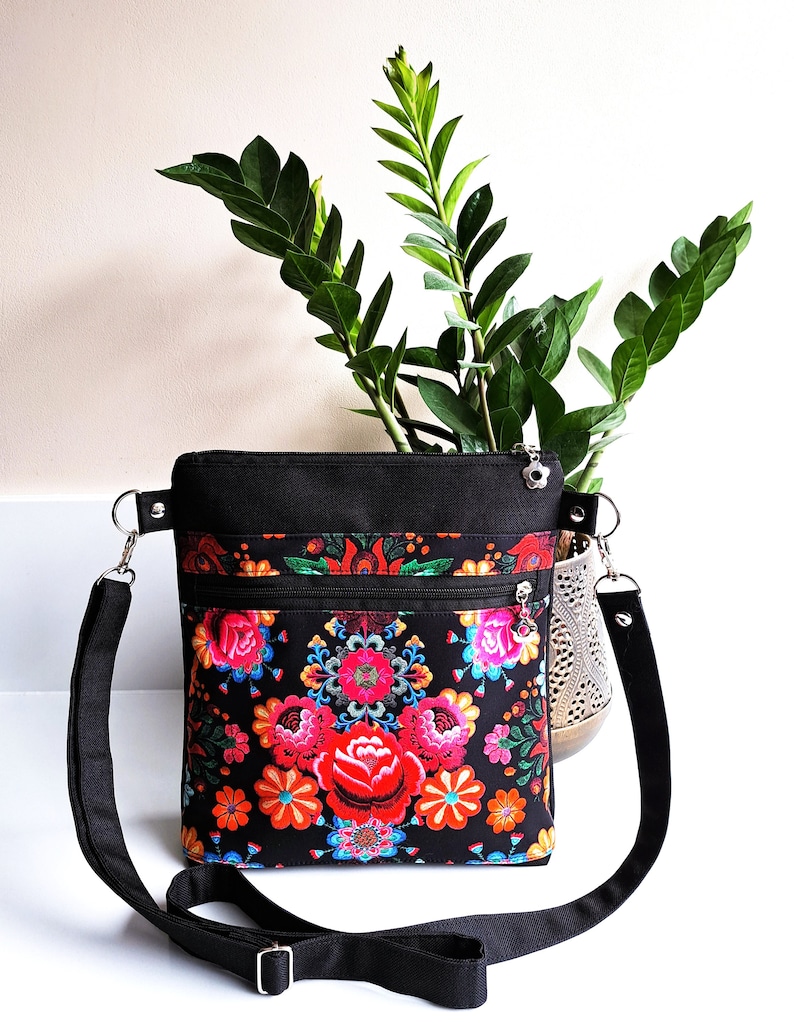 Pu&ograve; includere: Una borsa a tracolla nera con una stampa floreale con fiori rossi, rosa, arancioni, gialli, blu e verdi. La borsa ha una chiusura a zip e una tracolla regolabile.