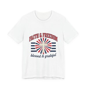 Puede incluir: Una camiseta blanca de manga corta con un diseño gráfico que incluye el texto FAITH & FREEDOM en rojo, una cruz beige central, un patrón de rayos de sol rojo y azul, rayas horizontales azules y el texto blessed & grateful en azul oscuro.