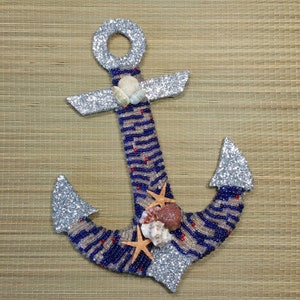 May include: A decorative anchor with a nautical theme. The anchor is embellished with blue and silver beads, seashells, and starfish. The anchor's top and flukes are covered in silver glitter. The anchor is a decorative item.