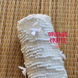 May include: White beaded bracelet with small white doves attached. The bracelet is made of white beads and has a textured surface. The doves are attached to the bracelet with a clear adhesive. The bracelet is on a woven brown background.