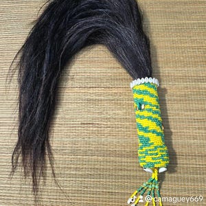 May include: A ceremonial item featuring a long, dark, flowing tail attached to a beaded handle. The handle is adorned with yellow and green beads in a zigzag pattern, and the base is decorated with small seashells. The item is likely used for cultural or spiritual purposes.