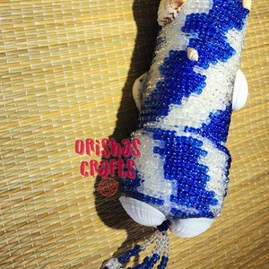 May include: A blue and white beaded cylinder with seashells attached. The cylinder has a fringe of blue and white beads hanging from the bottom. The cylinder is on a woven mat. The text "ORISHAS CRAFTS" is visible in the image.