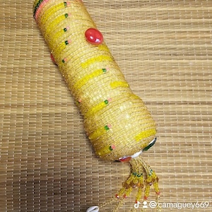 May include: A cylindrical, yellow beaded object with red accents and a beaded tassel with seashells. The object has a woven appearance and is decorated with yellow and pink stripes. The object is on a woven mat.