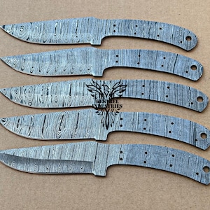 May include: Five Damascus steel knife blades with a wood grain pattern are arranged on a tan surface. The blades have a curved shape with a pointed tip and a handle section with pre-drilled holes. The text "Kestrel Industries" is visible on one of the blades.