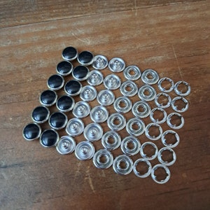 May include: A set of silver-toned snap fasteners, with black plastic buttons. The fasteners are arranged in a triangular shape, with the buttons at the top.