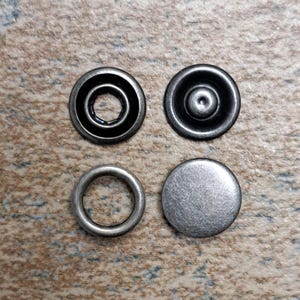 May include: Four antique silver-colored snap fasteners. Two are complete snaps, one is a ring, and the other is a flat disc. The fasteners are arranged in a square on a textured, neutral-toned surface.