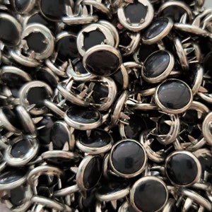 May include: A large pile of black and silver metal tacks with sharp points. The tacks have a round, flat head with a black center and a silver metal rim.