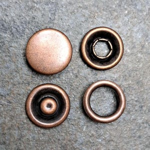 May include: Four antique copper-colored snap fasteners are arranged on a textured surface. The components include a flat, round cap, a socket with a ring of small metal pieces, a stud with a central post, and a ring.