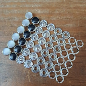 May include: A collection of silver snap fasteners and decorative buttons with black and pearlescent tops, arranged on a wooden surface. The fasteners are designed for clothing or craft projects.