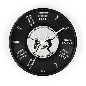 May include: Black vinyl record clock with white hands and a white silhouette of a couple dancing. The clock face has the text "We're gonna rock around the clock tonight" and the numbers 1 through 12 with the text "O'clock ROCK" at the 8, 4, and 12 o'clock positions.