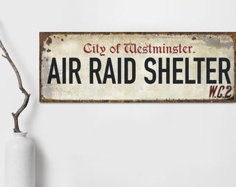 May include: A vintage-style metal sign with a white background and red and black text. The sign reads "City of Westminster. Air Raid Shelter W.C.2."