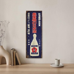 Könnte beinhalten: Ein Schild im Vintage-Stil, das japanischen Sake bewirbt. Das Schild ist blau mit weißen und roten Buchstaben. Es zeigt eine Flasche Sake mit einem roten Blumendesign und den Text "最高級の日本酒", der mit "Sake höchster Qualität" übersetzt wird.