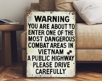 May include: A white sign with black text that reads "WARNING YOU ARE ABOUT TO ENTER ONE OF THE MOST DANGEROUS COMBAT AREAS IN VIETNAM A PUBLIC HIGHWAY PLEASE DRIVE CAREFULLY". The sign has a distressed, weathered look and appears to have bullet holes.