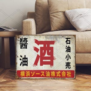 以下が含まれることがあります： 黒と赤の日本語テキストが入った、長方形の風化した金属看板。看板には日本語で「醤油」と「油」と書かれており、下部には赤い長方形にテキストが追加されています。看板は使い古されたヴィンテージの外観をしています。