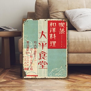 Op de afbeelding: Vintage metalen bord met Japanse tekst. Het bord heeft een rood en blauwgroen kleurenschema met Japanse karakters en een visillustratie. Het bord is verouderd met roest en slijtage.