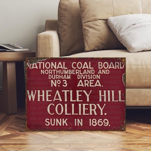 Puede incluir: Un letrero metálico rectangular desgastado, con fondo rojo y letras blancas. El letrero dice "NATIONAL COAL BOARD" y "WHEATLEY HILL COLLIERY. SUNK IN 1869." El letrero está ligeramente deteriorado.