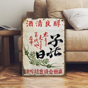 以下が含まれることがあります： 赤と黒の日本語の文字が入った、長方形のヴィンテージスタイルの金属看板。看板は、錆や摩耗が見られる、使い古された外観をしています。緑色の竹の小枝が下部にあります。看板はベージュのソファに立てかけられています。