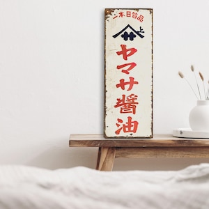 以下が含まれることがあります： 白い背景に赤い文字で書かれた、日本の古い看板。看板には日本語で「山井油」と「日本製品」と書かれている。看板は金属製で、素朴な外観をしている。