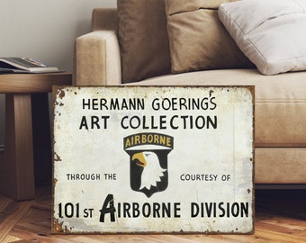 May include: A white metal sign with the text "Hermann Goering's Art Collection Through The 101st Airborne Division" and a black and white image of an eagle with the word "Airborne" above it.
