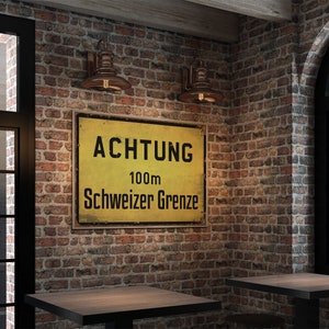 May include: A yellow and black sign with the text "ACHTUNG 100m Schweizer Grenze" on a brick wall with two copper wall sconces. The sign is mounted on the wall above two wooden tables.