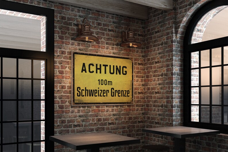 May include: A yellow and black sign with the text "ACHTUNG 100m Schweizer Grenze" on a brick wall with two copper wall sconces. The sign is mounted on the wall above two wooden tables.