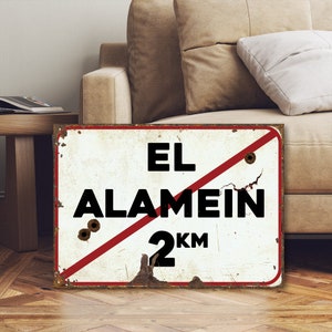 Peut inclure: Un panneau en métal blanc avec une ligne diagonale rouge et du texte noir qui dit "EL ALAMEIN 2 KM". Le panneau a un aspect vieilli et vintage avec de la rouille et des trous de balle visibles.