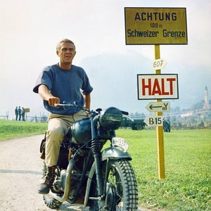 May include: A man in a blue shirt rides a black motorcycle on a dirt road. He is wearing brown pants and brown boots. A yellow sign with the text "ACHTUNG 100 m Schweizer Grenze" is visible in the background.