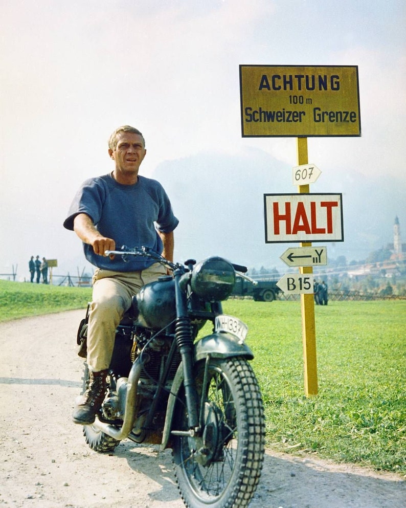May include: A man in a blue shirt rides a black motorcycle on a dirt road. He is wearing brown pants and brown boots. A yellow sign with the text "ACHTUNG 100 m Schweizer Grenze" is visible in the background.