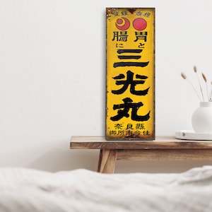 Op de afbeelding: Een vintage bord in geel en zwart met Japanse tekst die "Voor de huid, licht en helder." luidt. Het bord staat op een houten plank met een witte vaas en gedroogde bloemen.