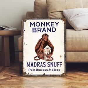 May include: Vintage tin sign advertising Monkey Brand Madras Snuff. The sign features a cartoon monkey sitting on a can of snuff, with the text "Monkey Brand Madras Snuff Post Box 555, Madras" in blue lettering.