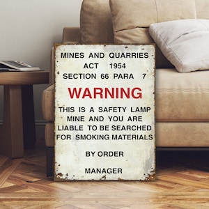 May include: A vintage-style warning sign with the text "Mines and Quarries Act 1954 Section 66 Para 7 Warning This is a safety lamp mine and you are liable to be searched for smoking materials By Order Manager". The sign is white with red text and is slightly weathered.