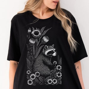 May include: Black t-shirt with a white graphic print of a raccoon surrounded by flowers. The raccoon is looking to the right and has a white nose and white markings around its eyes.