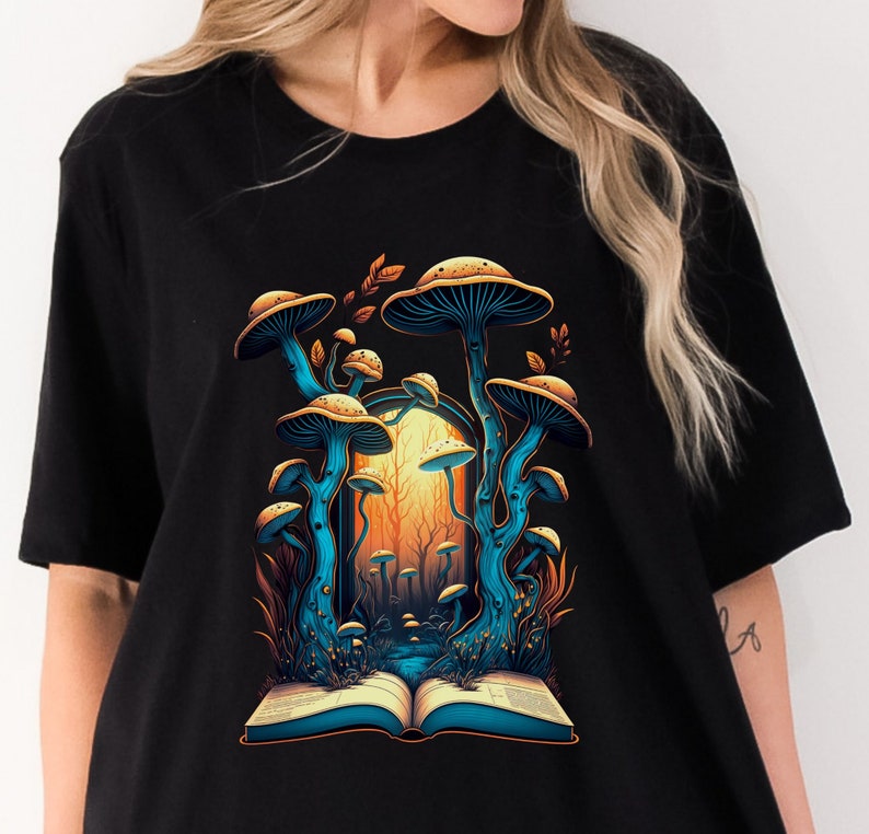 May include: Black t-shirt featuring a vibrant illustration of blue and tan mushrooms with a forest backdrop. The design includes an open book at the base of the mushrooms, creating a whimsical and nature-inspired aesthetic.
