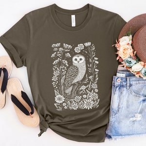 May include: A dark green t-shirt with a white print of an owl surrounded by flowers. The owl is facing forward with its wings spread out. The flowers are in bloom and have white petals.