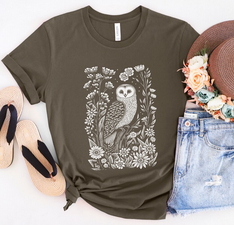 May include: A dark green t-shirt with a white print of an owl surrounded by flowers. The owl is facing forward with its wings spread out. The flowers are in bloom and have white petals.