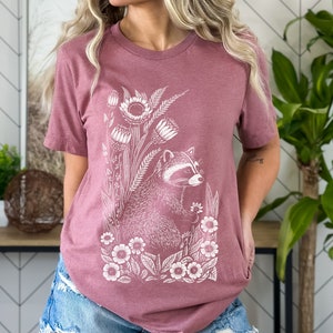 May include: A mauve t-shirt with a white graphic of a raccoon surrounded by flowers and leaves.