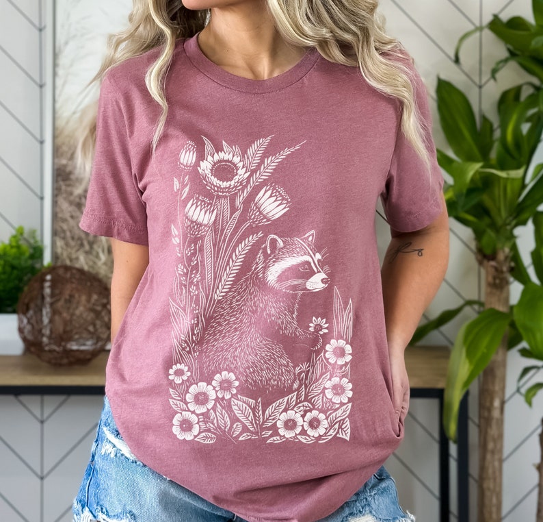 May include: A mauve t-shirt with a white graphic of a raccoon surrounded by flowers and leaves.