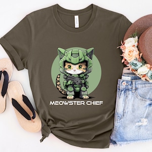 Puede incluir: Camiseta verde oliva con un gato de dibujos animados con casco y armadura militar verde, sosteniendo un arma. El texto "MEOWSTER CHIEF" est&aacute; impreso debajo de la ilustraci&oacute;n del gato. La camiseta se muestra con un sombrero de paja y pantalones cortos de mezclilla.