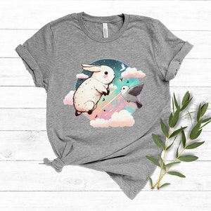 May include: A gray t-shirt with a white rabbit and a bird flying in a circle of clouds. The rabbit is jumping and the bird is flying towards the rabbit. The background is a pastel blue and pink.