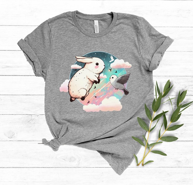 May include: A gray t-shirt with a white rabbit and a bird flying in a circle of clouds. The rabbit is jumping and the bird is flying towards the rabbit. The background is a pastel blue and pink.