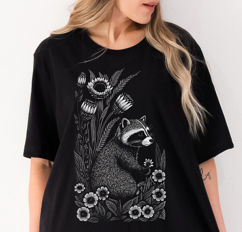 May include: Black t-shirt with a white graphic print of a raccoon surrounded by flowers. The raccoon is looking to the right and has a white nose and white markings around its eyes.