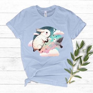 May include: A light blue t-shirt with a graphic of a white rabbit and a gray bird flying in a circle of clouds with a pink, blue, and green starry sky.