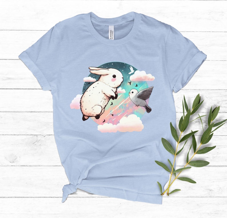 May include: A light blue t-shirt with a graphic of a white rabbit and a gray bird flying in a circle of clouds with a pink, blue, and green starry sky.