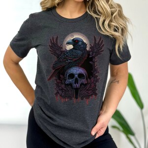 May include: A dark gray t-shirt with a graphic print of a black raven perched on a skull with red eyes. The raven is surrounded by a moon and floral designs.