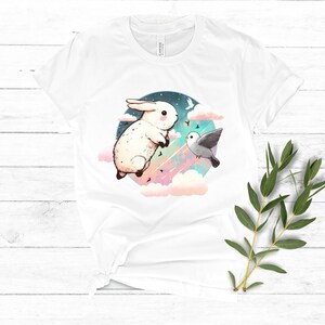 May include: White t-shirt with a graphic of a white rabbit and a gray bird flying in a colorful sky with clouds.