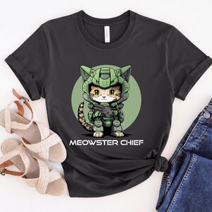 Puede incluir: Camiseta gris oscuro con un gato de dibujos animados con casco y armadura militar verde, sosteniendo un arma. El gato est&aacute; sobre un c&iacute;rculo verde, con el texto "MEOWSTER CHIEF" debajo.
