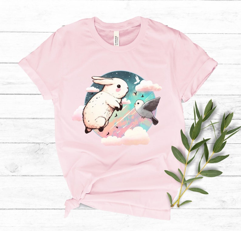 May include: A pink t-shirt with a white rabbit and a bird flying in the clouds. The rabbit is jumping and the bird is flying towards the rabbit. The background is a blue sky with stars and clouds.